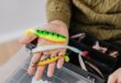 Close-up of colorful fishing lures in a hand, next to a fishing tackle box.