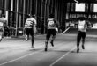 Back view of athletes sprinting on an indoor track, captured in grayscale.
