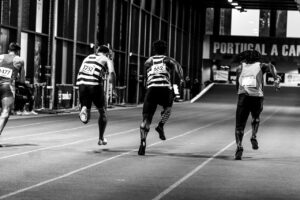 Back view of athletes sprinting on an indoor track, captured in grayscale.