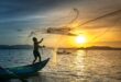 A fisherman skillfully casts a net at sunrise, silhouetted against a golden sky over a calm lake.